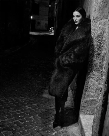 Model in dark oversized fur coat leaning against a stone wall in a dark city alley at night