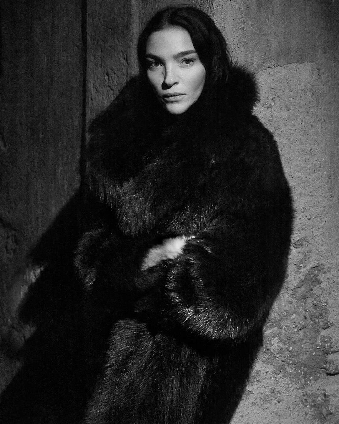 Monochrome portrait of Mariacarla Boscono in a dark textured fur coat posing against a rough stone wall