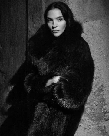 Monochrome portrait of Mariacarla Boscono in a dark textured fur coat posing against a rough stone wall