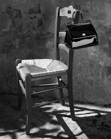 Black patent leather handbag hanging from a rustic wooden chair against a weathered stone wall in monochrome