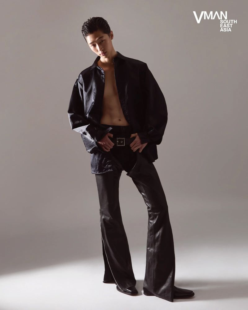 Model in unbuttoned black shirt and leather pants standing against a plain light grey studio backdrop.