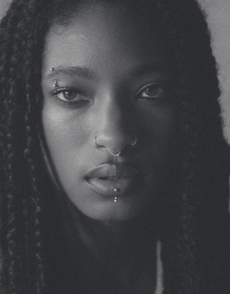 Close-up black and white portrait of a young adult female with dark braided hair, visible facial piercings, and a neutral expression.