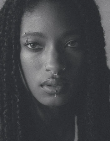 Close-up black and white portrait of a young adult female with dark braided hair, visible facial piercings, and a neutral expression.