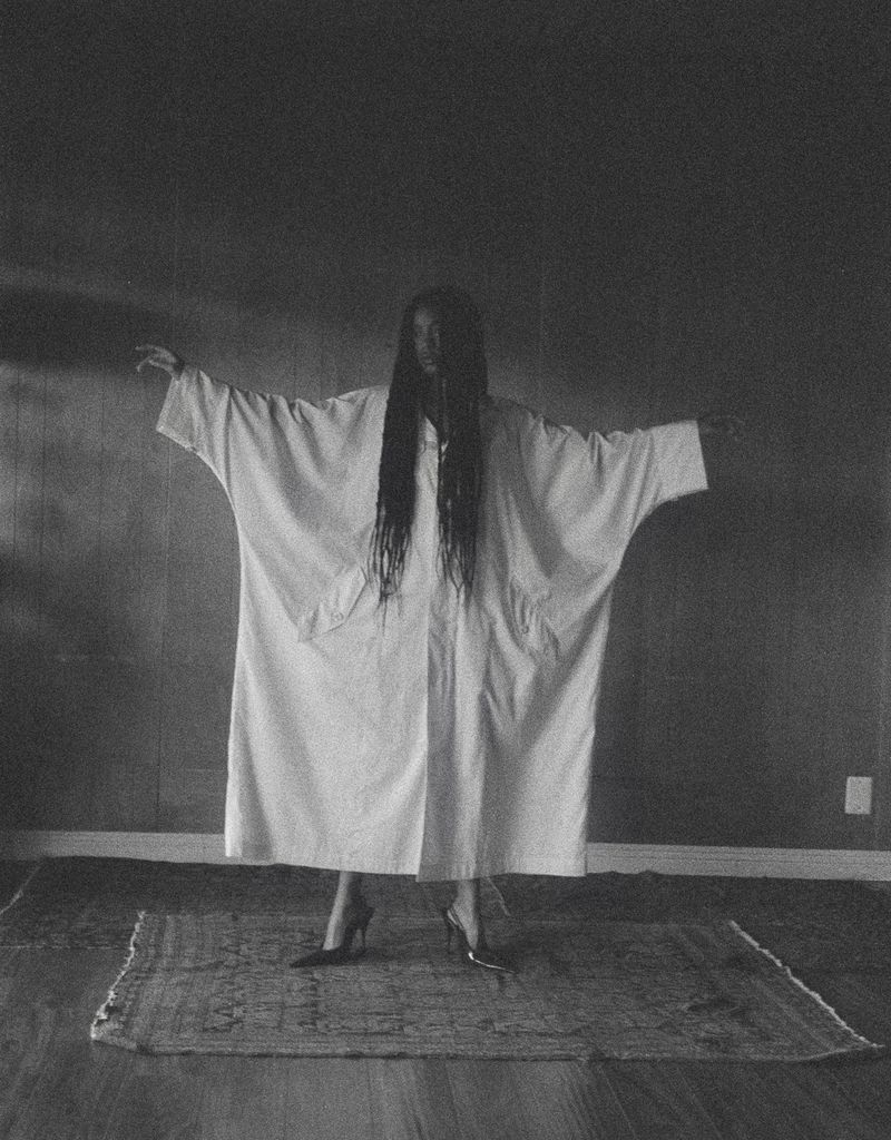 Person in white robe stands on a rug with arms outstretched, black hair covering their face in a dimly lit indoor space.