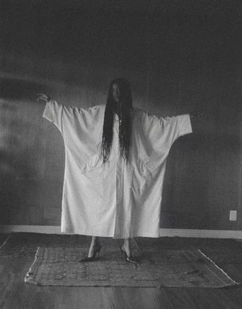Person in white robe stands on a rug with arms outstretched, black hair covering their face in a dimly lit indoor space.