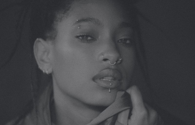 Close-up black and white portrait of an adult female with multiple facial piercings in an indoor setting with dark lighting.