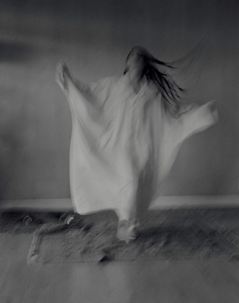 Motion-blurred monochrome image of an adult with long hair, in a flowing white garment, raising arms slightly, walking on textured ground.