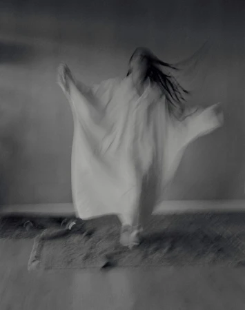 Motion-blurred monochrome image of an adult with long hair, in a flowing white garment, raising arms slightly, walking on textured ground.