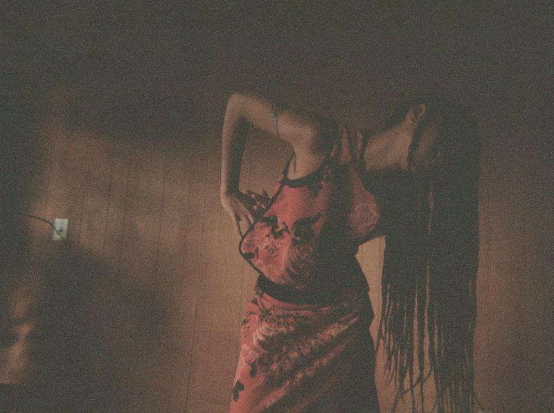 Adult female in a pink floral top and matching skirt poses in a room with dark lighting, and a wall with an electrical outlet.