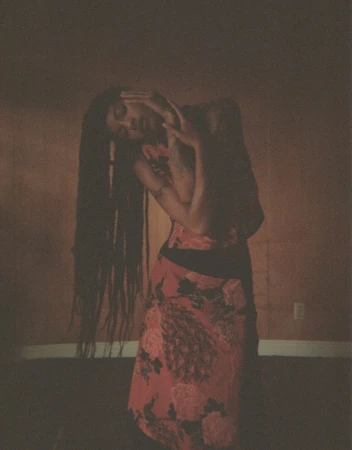 Woman with long dreadlocks in floral dress poses indoors against warm-toned walls, expressive hands framing her face in soft lighting.