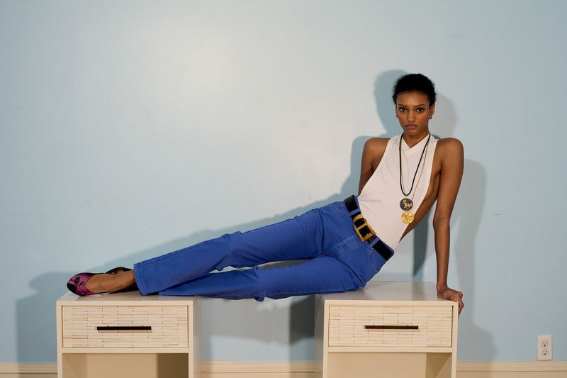 Model wearing blue pants and white halter top reclining across two white nightstands against light blue wall.
