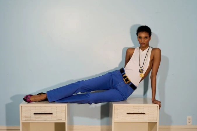 Model wearing blue pants and white halter top reclining across two white nightstands against light blue wall.