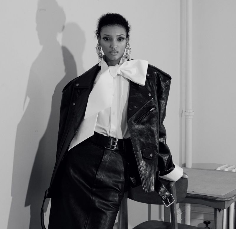Model in a black leather jacket and white shirt with a large bow standing against a plain wall