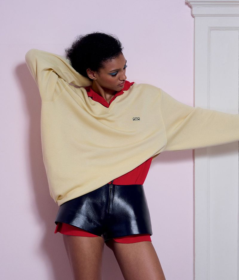 Model wearing a yellow oversized sweater and dark leather shorts against a light pink wall.