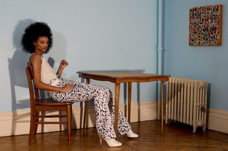Model wearing patterned pants and a light tank top seated on a wooden chair indoors against a pale blue wall.