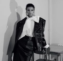 Model in a black leather jacket and white shirt with a large bow standing against a plain wall