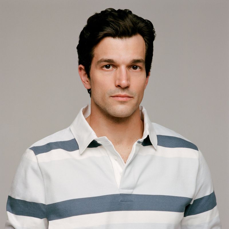 Portrait of Paul Anthony Kelly wearing a white and blue striped polo shirt against a plain grey background