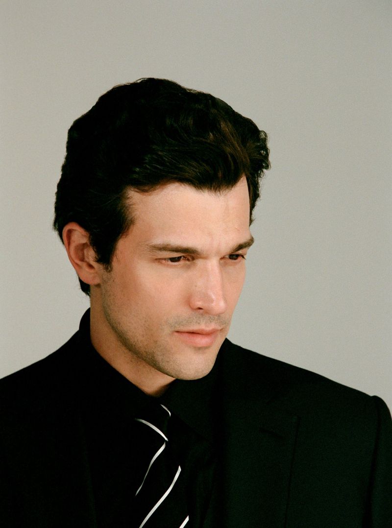 Portrait of a man wearing a black suit and striped tie against a plain grey backdrop.