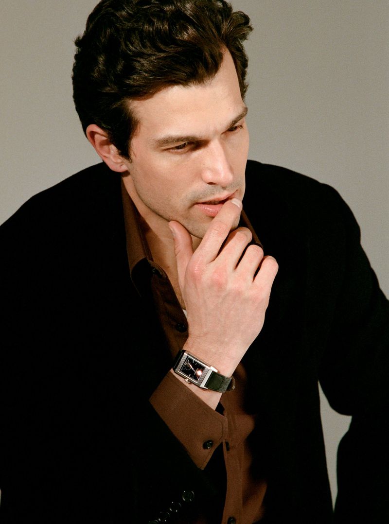 Man with dark hair wearing a brown shirt and black jacket with a watch on his wrist against a grey backdrop