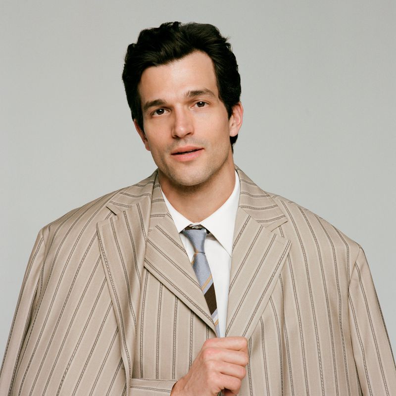 Portrait of a man in an oversized pinstripe suit and tie, gazing directly at the camera against a plain, light grey backdrop.