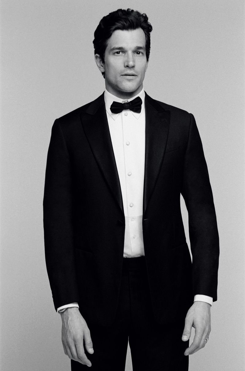 Man wearing a black tuxedo with a white shirt and black bow tie against a seamless grey studio background.