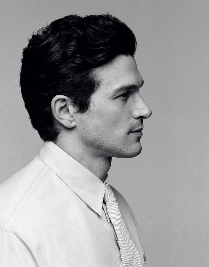 Profile portrait of a man with dark wavy hair wearing a collared button-down shirt and tie against a plain grey backdrop.