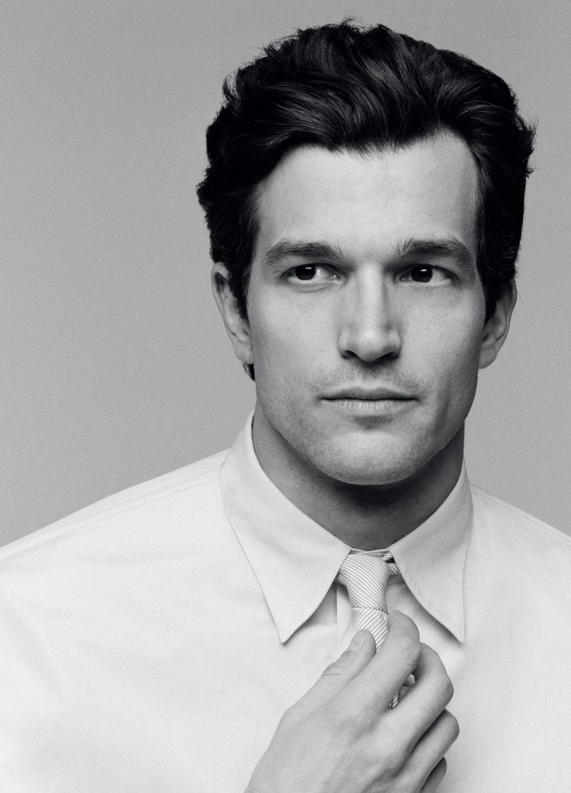 Portrait of Paul Anthony Kelly adjusting his tie wearing a white dress shirt against a plain grey studio background