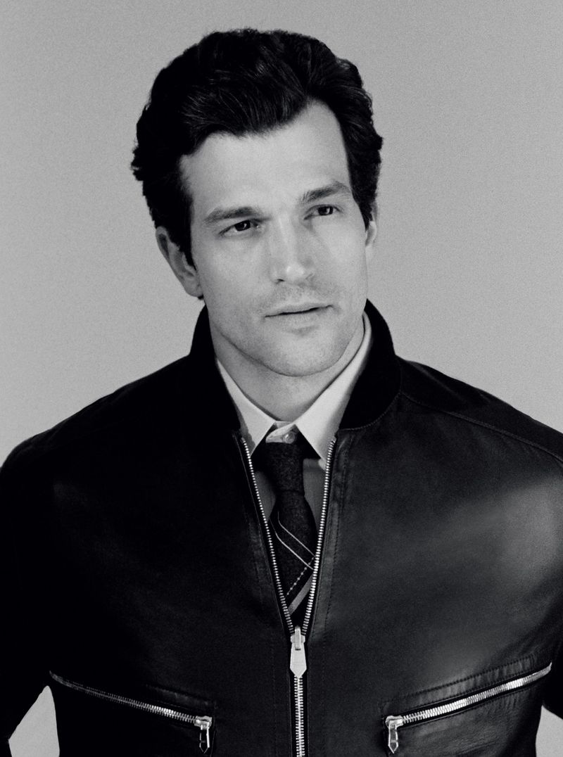 Black and white portrait of a male model wearing a dark leather bomber jacket, collared shirt, and striped tie against a neutral background.