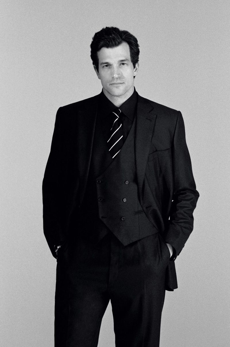 Model Paul Anthony Kelly in a black three-piece suit and striped tie against a plain grey seamless background.