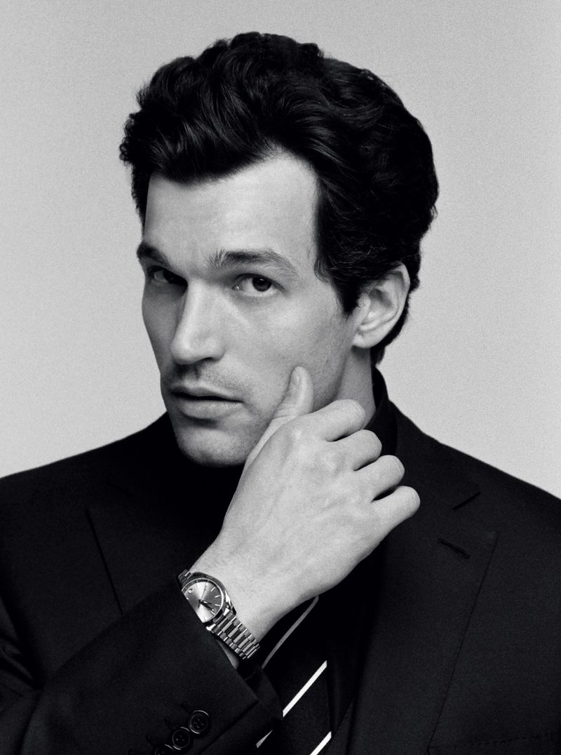 Black and white portrait of a man wearing a dark suit jacket, tie, and metal wristwatch against a plain light background