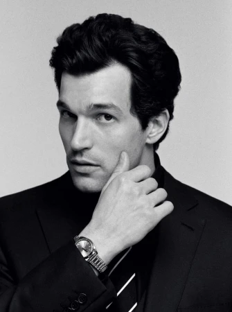 Black and white portrait of a man wearing a dark suit jacket, tie, and metal wristwatch against a plain light background