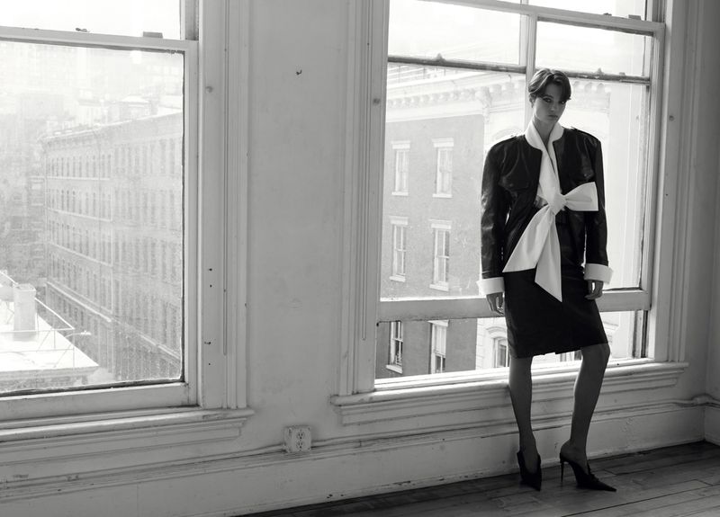 Model standing in front of large windows wearing a dark jacket with a large white bow and dark skirt.