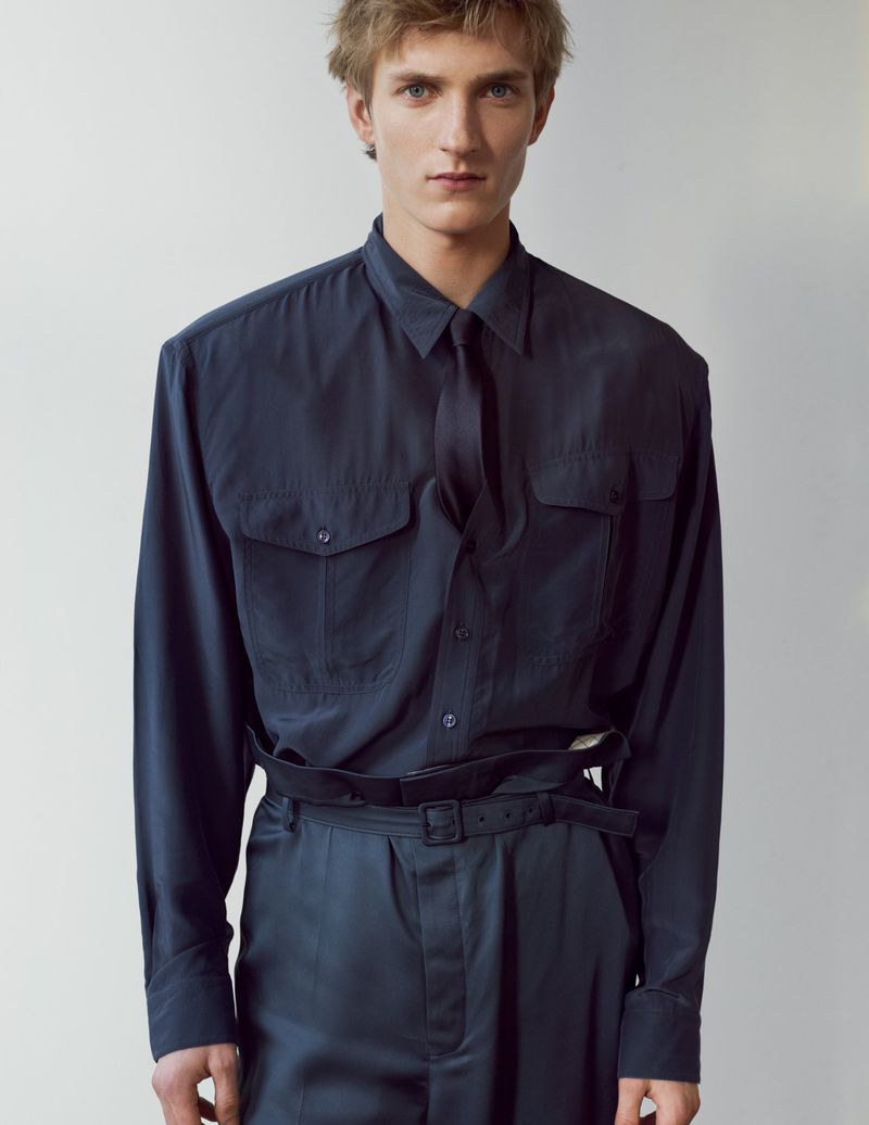Young male model wearing a dark button-down shirt and tie against a plain light wall
