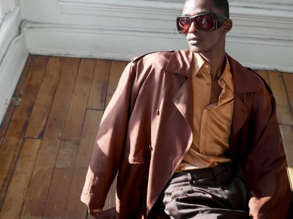 Man wearing oversized sunglasses, a gold satin shirt, and a brown coat seated on a wooden floor.