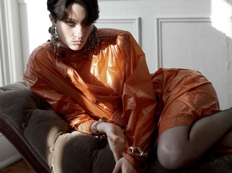 Model in shiny orange dress and large earrings reclining on a tufted chaise lounge in natural sunlight