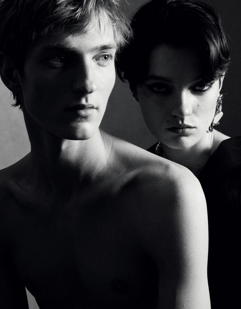 Monochrome portrait of a shirtless man and a woman in dark attire standing closely together in studio lighting.