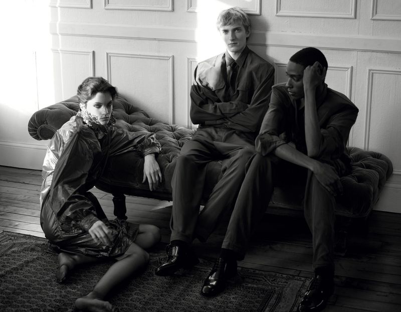 Three models posed in an interior setting with a tufted chaise lounge and wood panel walls in a black and white fashion image.
