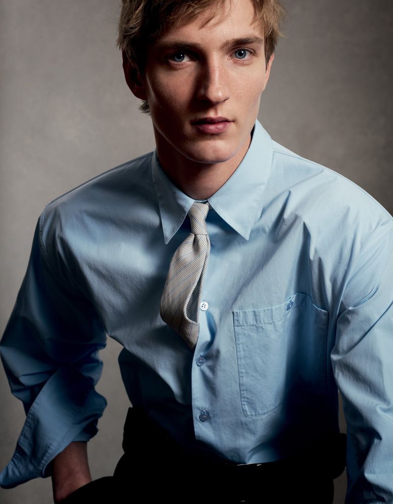 Close-up portrait of a young man wearing a light blue button-down shirt and a grey tie against a neutral grey background.