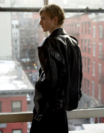 Young man in a black leather jacket standing by a window looking out at a city street