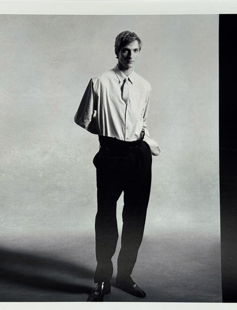 Monochrome portrait of a male model wearing a light button-down shirt and dark trousers against a neutral textured studio background.