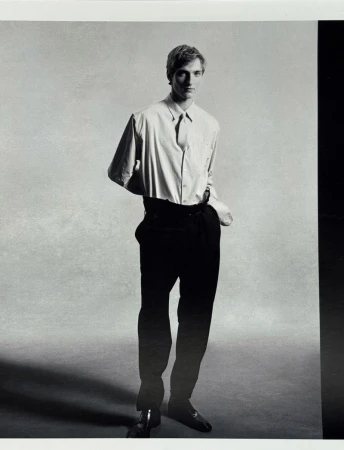 Monochrome portrait of a male model wearing a light button-down shirt and dark trousers against a neutral textured studio background.