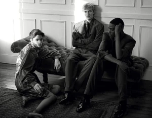 Three models posed in an interior setting with a tufted chaise lounge and wood panel walls in a black and white fashion image.