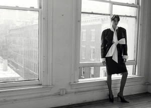 Model standing in front of large windows wearing a dark jacket with a large white bow and dark skirt.