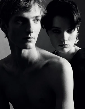 Monochrome portrait of a shirtless man and a woman in dark attire standing closely together in studio lighting.