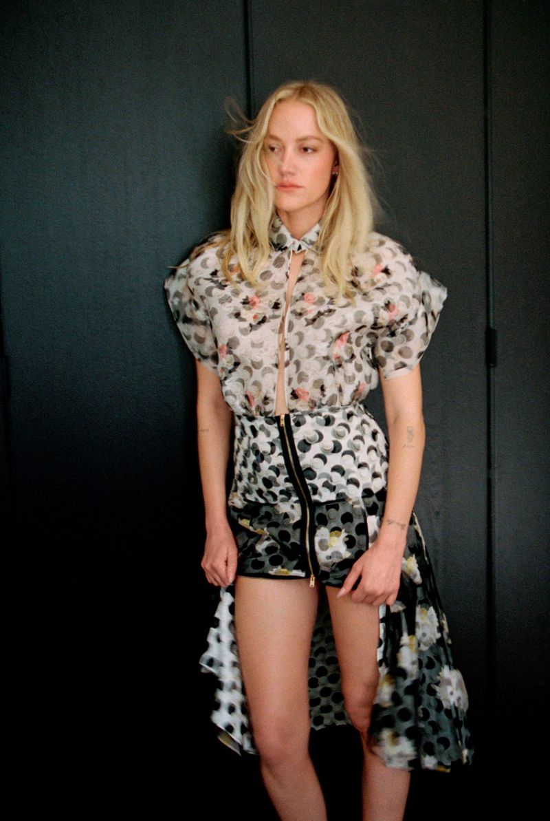 Maika Monroe wearing a patterned short sleeve top and mini skirt standing against a dark panelled wall.