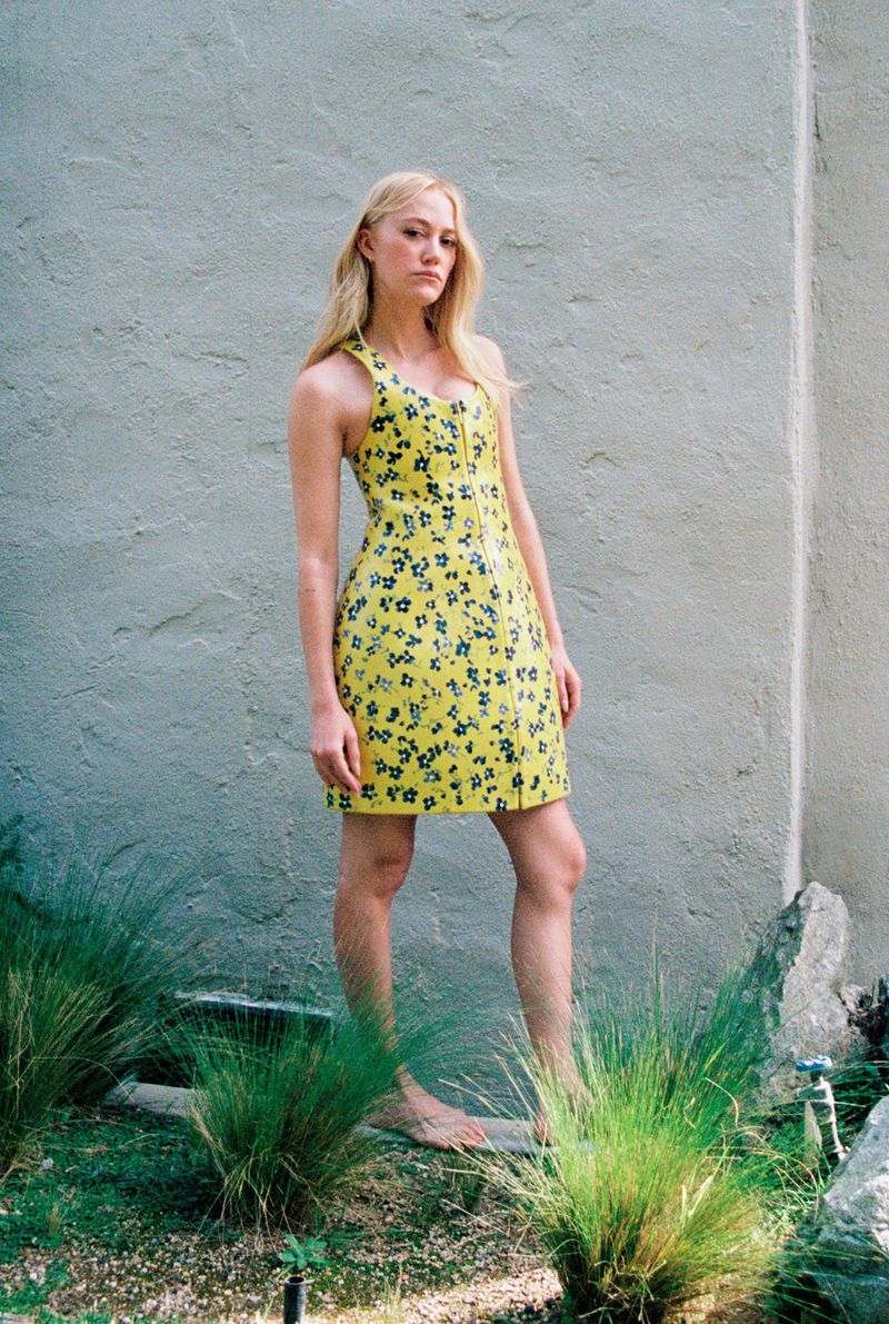 Maika Monroe wearing a yellow floral dress standing outdoors against a textured wall with foliage