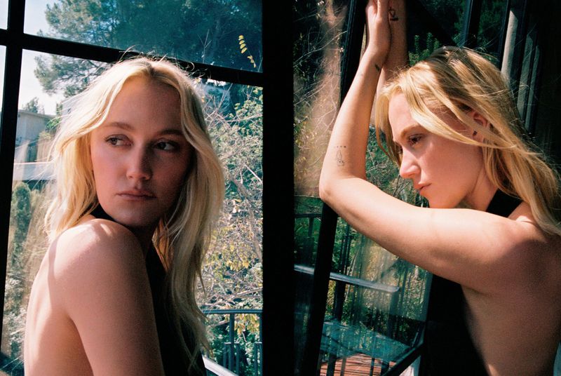 Maika Monroe posing indoors near a dark window frame with natural daylight illuminating her blonde hair