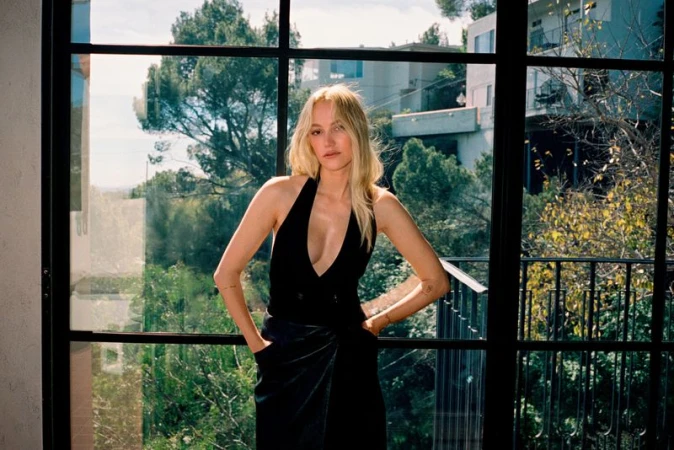 Maika Monroe posing indoors against large black-framed windows overlooking a green leafy exterior and residential buildings.