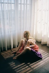 Maika Monroe seated on a wooden floor in front of sheer white window curtains, wearing a purple and pink dress.