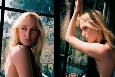 Maika Monroe posing indoors near a dark window frame with natural daylight illuminating her blonde hair